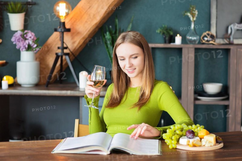 Young woman reading an interesting magazine at wooden table with fruits and wine on a weekend at home