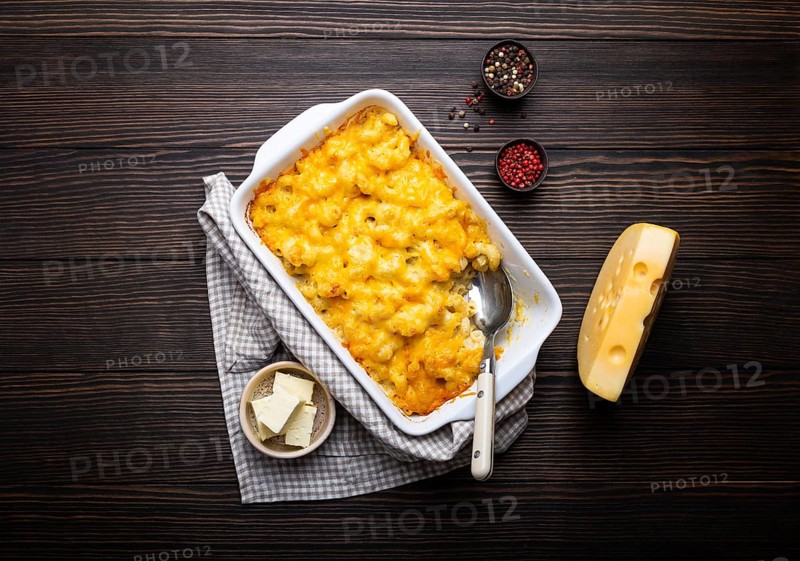 Close-up of baked mac and cheese in white casserole on rustic wooden background, with seasonings, butter, spoon and fork, top view. Pasta with cheesy sauce, American/English comfort food