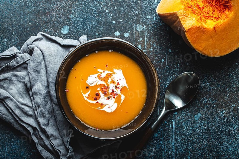 Homemade pumpkin cream soup served in brown ceramic bowl on dark stone background with spoon decorated with cut fresh pumpkin, top view. Traditional autumn comfort food