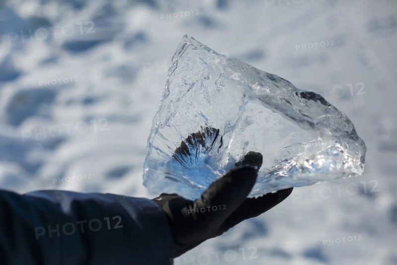 Happy smiling man wearing winter blue jacket and holding piece of transparent crushed ice cubes in his hands. Sun is shining through the sides of ice cubes. Floes look like diamonds