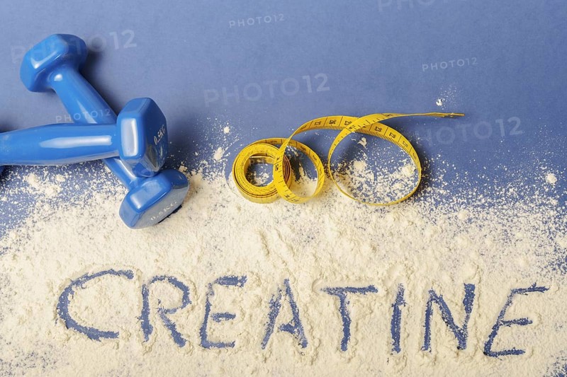 Word creatine written on creatine powder on blue background, with dumbbells and measuring tape