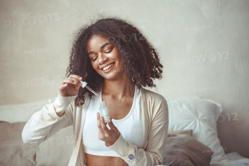 Beauty and skincare concept. Cropped photo of african american young female model holding bottle with moisturizing essence serum sitting against blurred modern apartment interior background