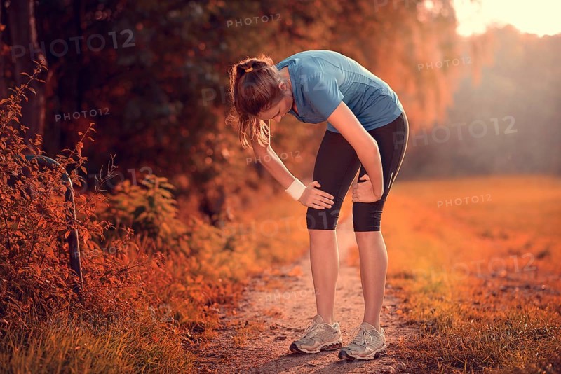 Young athletic woman taking a break from training standing resting her hands on her knees on a rural track through lush farmland in a health and fitness concept