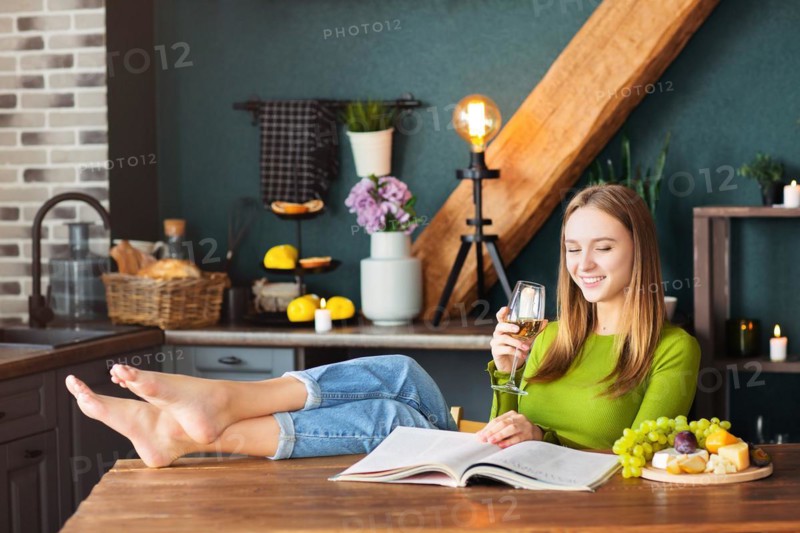 Young woman reading an interesting magazine at wooden table with fruits and wine on a weekend at home
