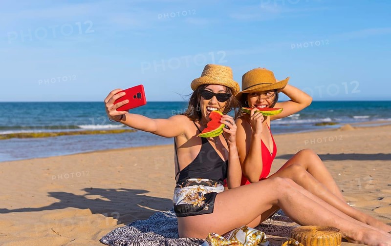 Friends on the beach in summer eating a watermelon with the sea in the background, taking a selfie and smiling