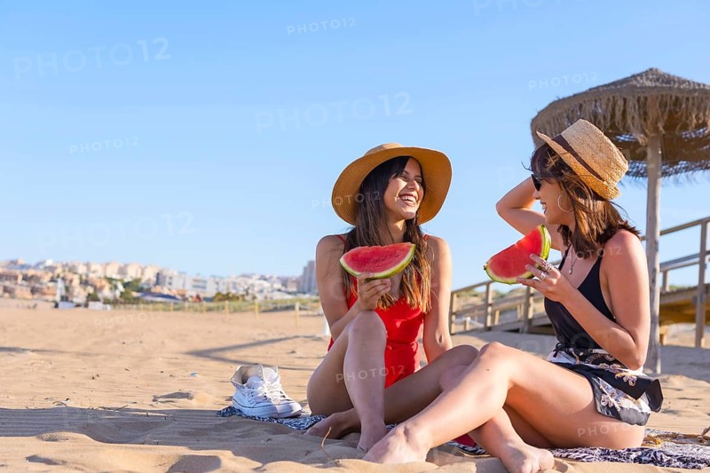 Female friends on vacation at the beach in summer eating a watermelon, sitting on the sand next to an umbrella