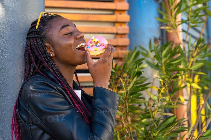Cheerful African woman with leather jacket biting a doughnut in the corner of street