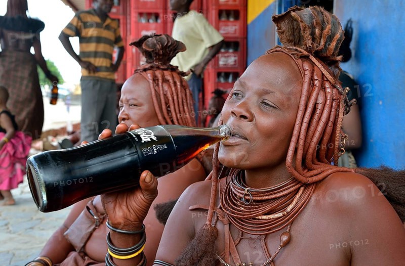 Traditionally dressed Himba woman drinking a coke in front of a bar, Opuwo, Kaokoland, Kunene, Namibia, Africa
