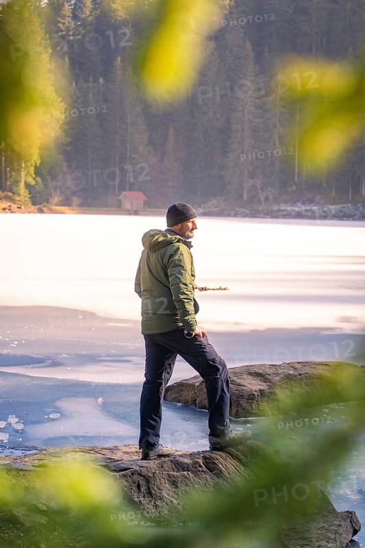 Person standing on the shore of a frozen lake, framed by rocky shore and forest, Glaswaldsee, Bad Rippoldsau-Schapbach, district of Wolfach, Black Forest, Germany, Europe