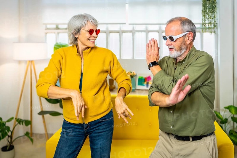 Frontal three quarter length photo of a caucasian married senior couple dancing wearing modern sunglasses at home