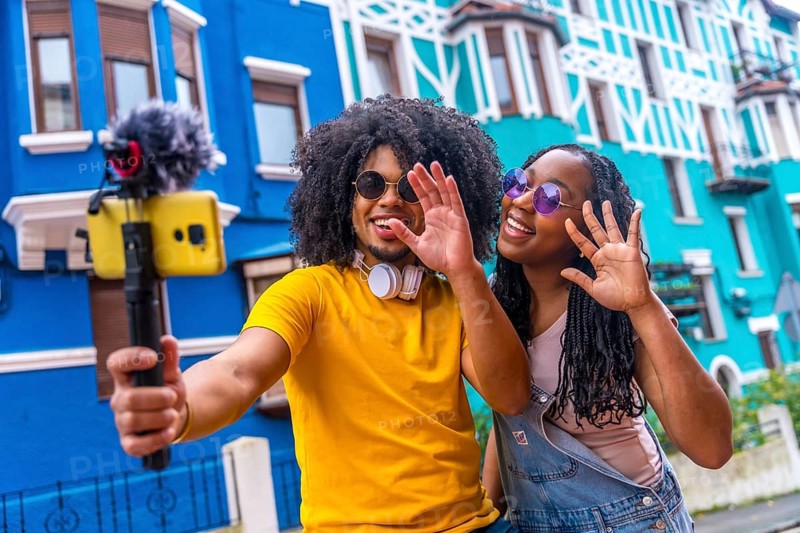 African couple broadcasting a life video waving at camera next to blue colorful house
