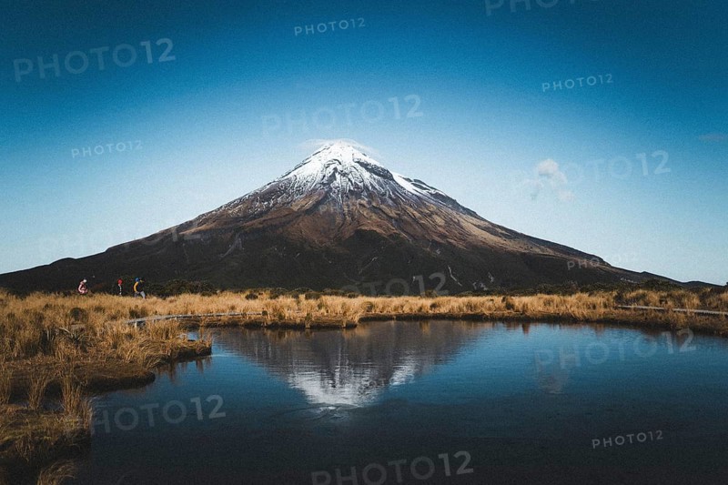 A snow-capped mountain is reflected in a still lake while people walk along the shore, Mount Taranaki, New Zealand, Oceania