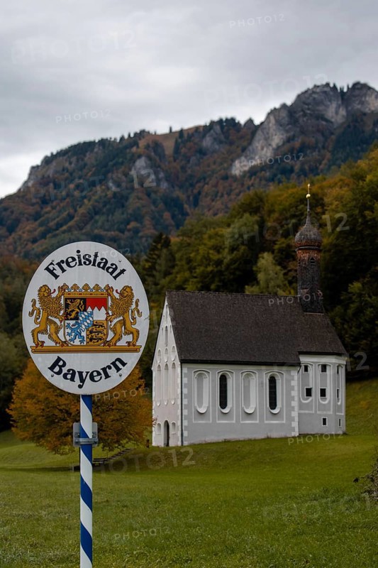 Free State of Bavaria sign with Nussdorf-Windshausen Kreuzkirche in the background, autumnal mountain landscape (Heuberg) with overcast sky, surrounded by trees and a green meadow, Nussdorf, Bavaria, Germany, Europe