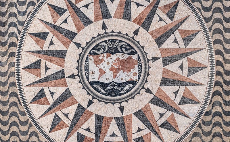 Aerial view of pavement map with routes of Portuguese explorations by Monument to Discoveries, Belem, Lisbon, Portugal, Europe. Antique map maritime motifs, compass, wind rose, image decorations, calcada patterns made of pale, black and red limestone, Europe