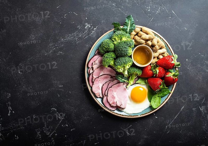 Ketogenic low carbs diet, top view, space for text. Plate on stone black background with keto foods: egg, meat, olive oil, broccoli, berries, nuts, seeds. Healthy fats, clean eating for weight loss
