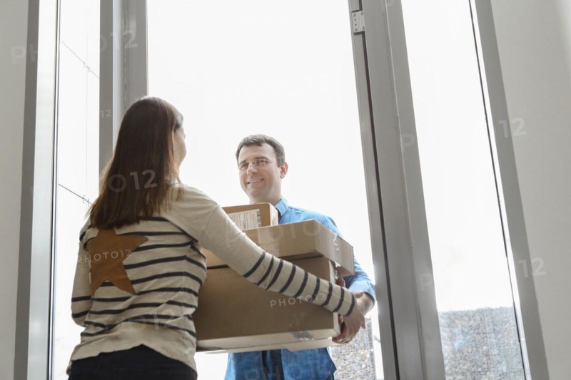 Fast and reliable service. Cheerful young courier giving a cardboard box to a young woman standing at the entrance to her apartment