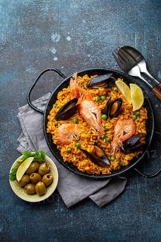 Classic dish of Spain, seafood paella in traditional pan on rustic blue concrete background top view. Spanish paella with shrimps, clamps, mussels, green peas and fresh lemon wedges from above