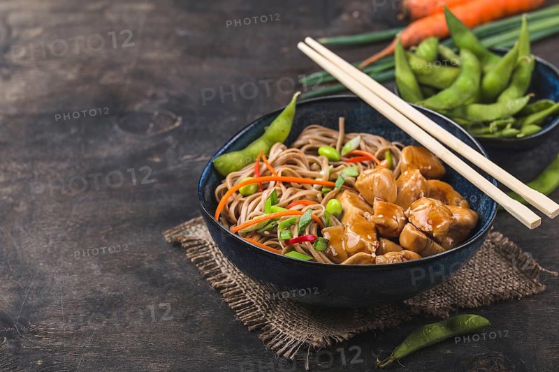 Asian noodles with chicken, vegetables in bowl, rustic wooden background. Space for text. Soba noodles, teriyaki chicken, edamame, chopsticks. Closeup. Asian style dinner. Chinese/Japanese noodles