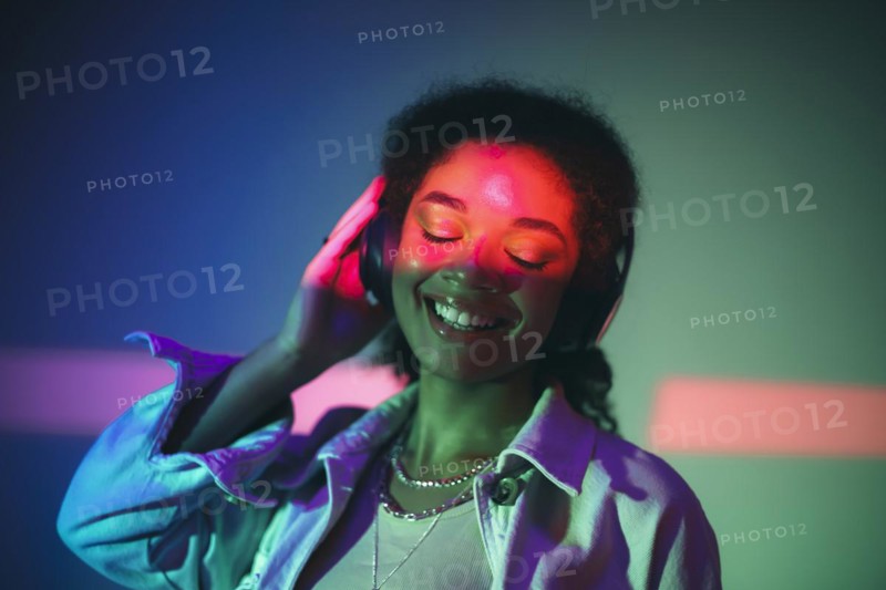 Young happy african american woman in black headphones listening to music, enjoying high quality sound while standing under purple neon glowing light in studio and smiling happily. Musicial equipment