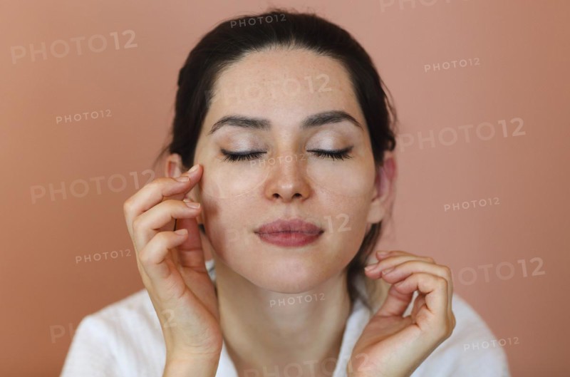 Positive young brunette female with eye patches touching her face and smiling during skin care routine