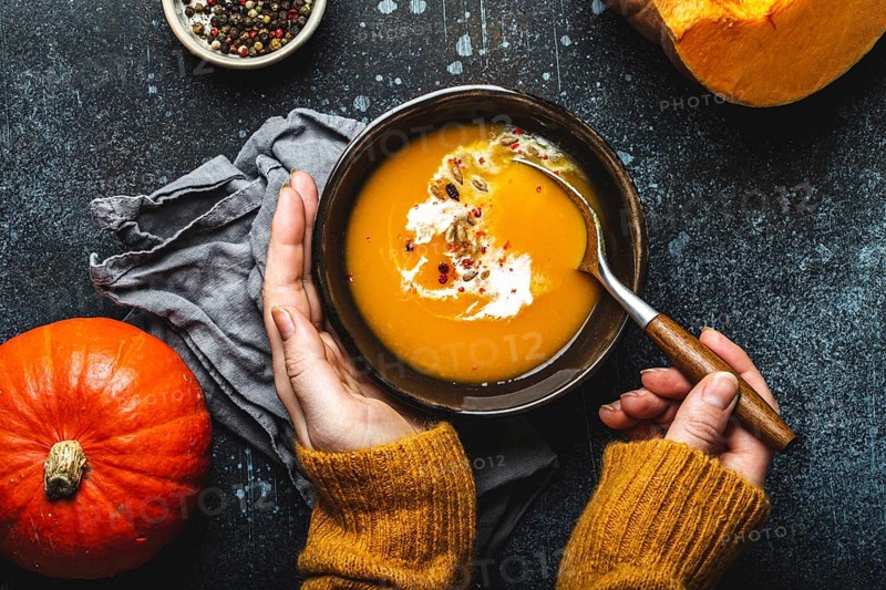 Woman in yellow knitted sweater eating a pumpkin cream soup on dark stone background with spoon decorated with cut fresh pumpkin, top view. Autumn cozy dinner concept