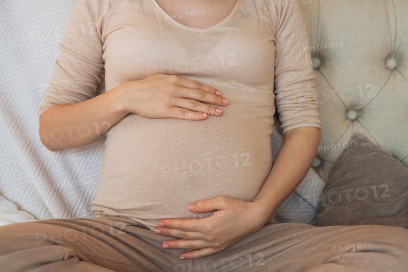 Cropped shot of pregnant woman holding belly while relaxing on couch at home, expectant mother sitting on sofa indoors and gently touching tummy. Pregnancy, maternity and prenatal period concept