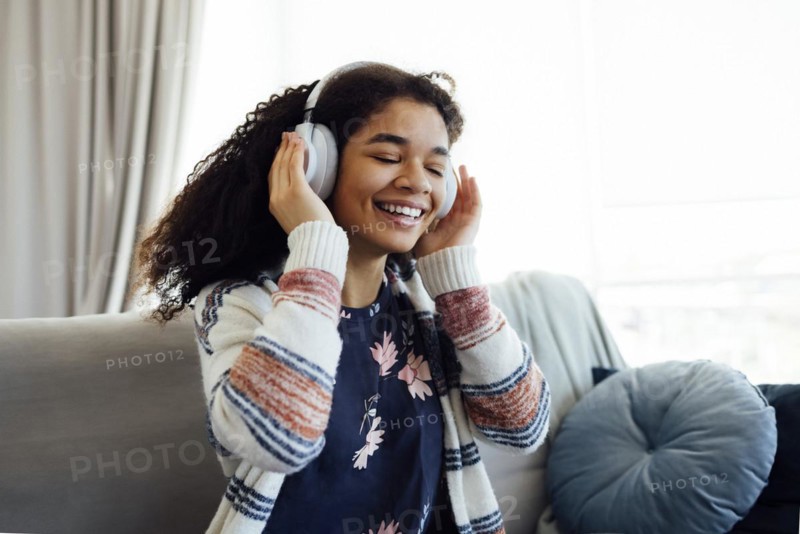African millennial woman in orange sweater wearing white wireless headphones, listening music, resting and lying on couch in living room. Girl spend lazy weekend at home