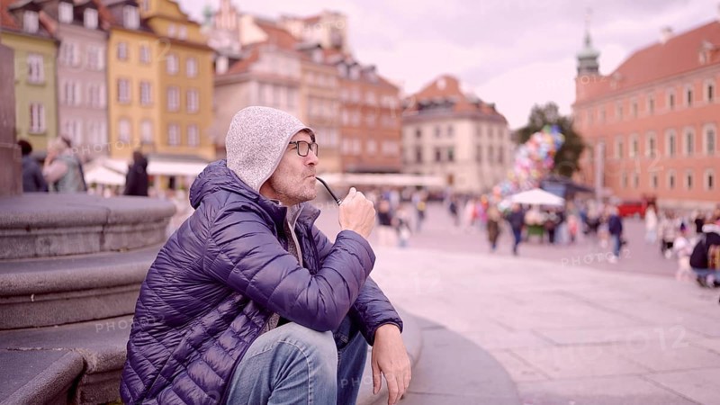 Portrait of adult man with glasses sitting in the hood on square and smoking a tobacco pipe in the Palace Square, Warsaw Old Town