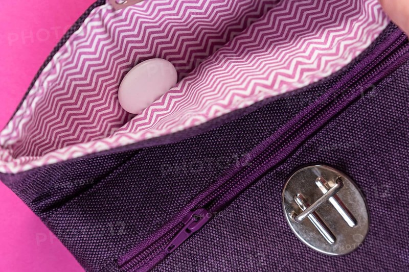 An Apple Air Tag lies in a purple clutch against a plain pink background