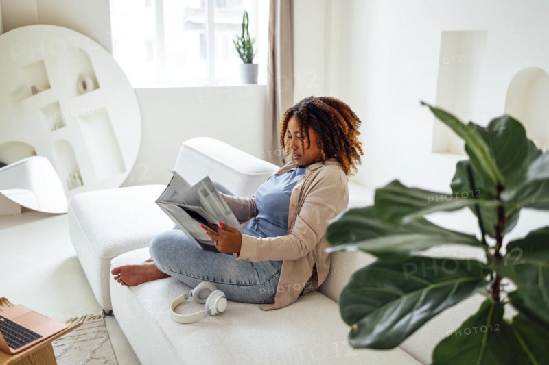 Photo of pretty happy young african woman sitting on sofa indoors at home while reading magazine or book