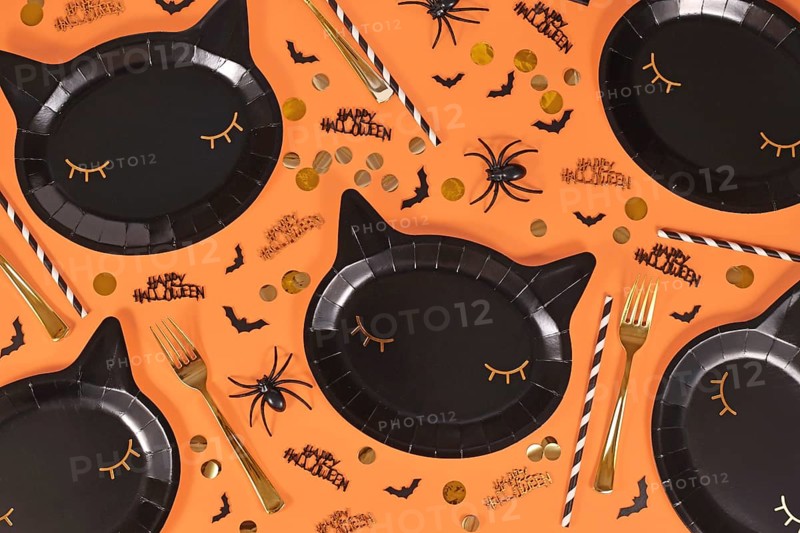 Black cat shaped Halloween party paper plates, bats and Happy Halloween text confetti on orange background