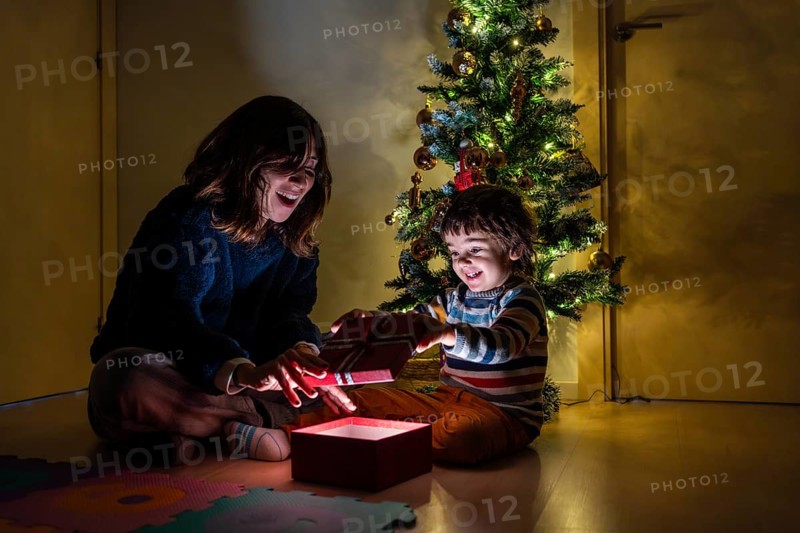 A woman and a child are opening a present in front of a Christmas tree. Scene is joyful and festive, as the two are sharing a special moment together during the holiday season
