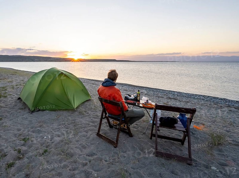 Camping at the lakeshore, young man sitting next to tent on camping chair during dinner, sunset at Issyk Kul Lake, Issyk Kul, Kyrgyzstan, Asia
