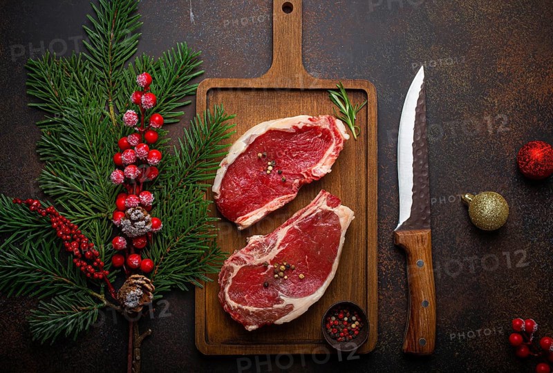 Two raw uncooked meat beef steaks on wooden cutting board with knife and seasonings on dark rustic background with Christmas festive decoration from above, food photography