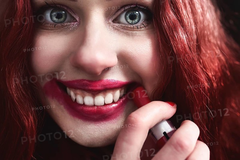 Portrait of crazy-looking teen girl with red hair she is smearing red lipstick on her face, horror concept. halloween time. Fear and nightmare. close view