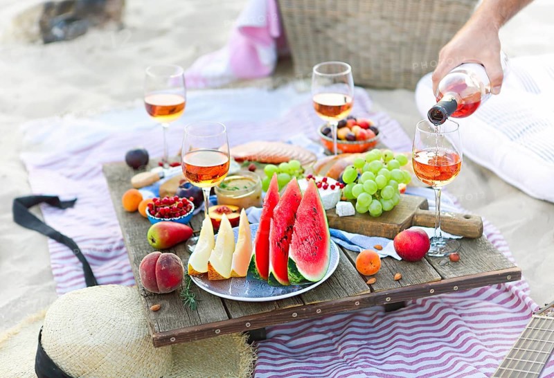 Crop male serving pink wine having picnic with delicious snacks and fruit on sandy beach