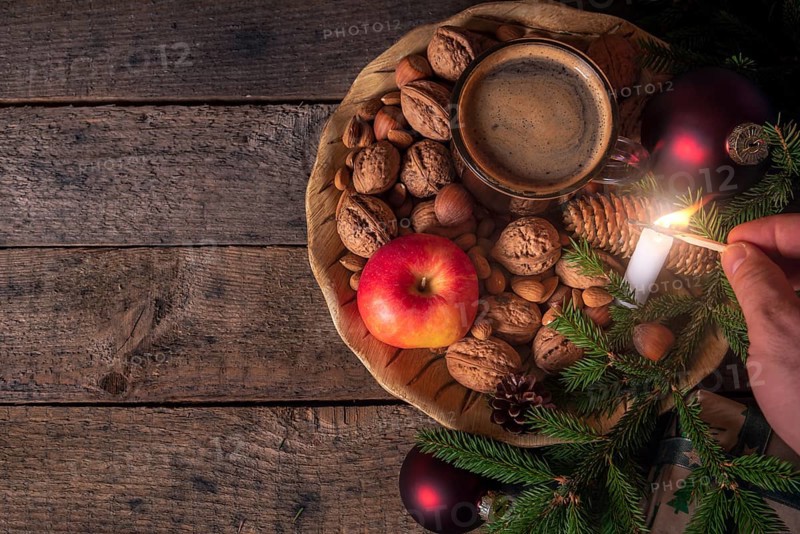 Christmas table with a wooden plate full with nuts, apples, a cup of coffee and green fir twigs, while a man lights up a candle. Low light image