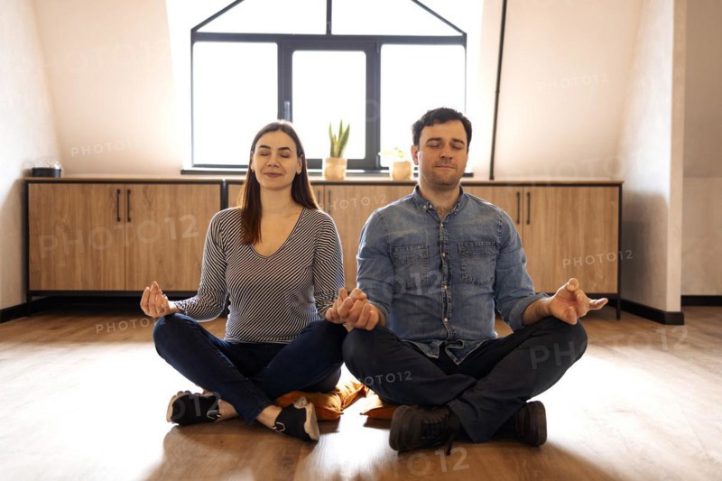 Peaceful adult couple in sportswear sitting on the floor in lotus pose with eyes closed, meditating together at home. Calm people, husband and wife, having yoga practice, relaxation concept