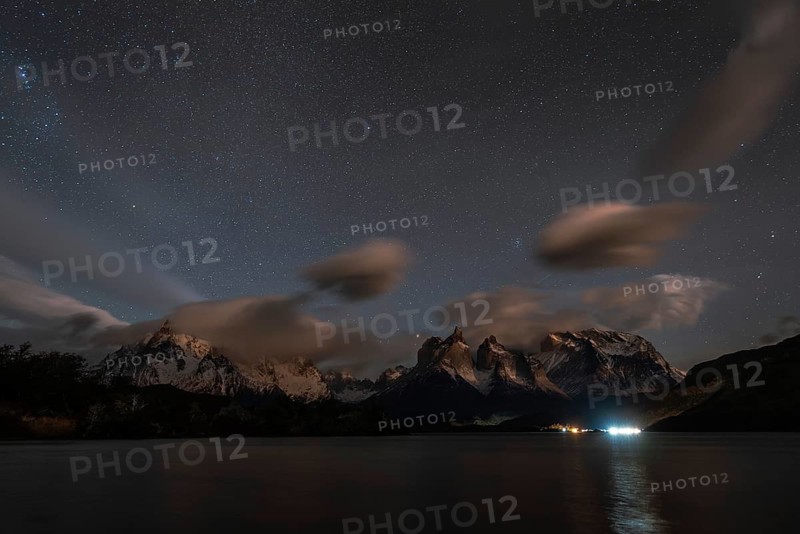 Night sky filled with stars over snow-capped mountains and a calm lake, with clouds and reflections creating a serene atmosphere, Torres Del Paine, Patagonia