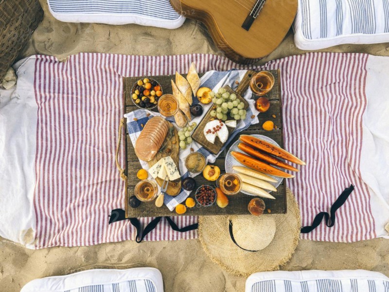 Various tasty food and drinks placed on wooden table during summer picnic on sandy beach at sunset