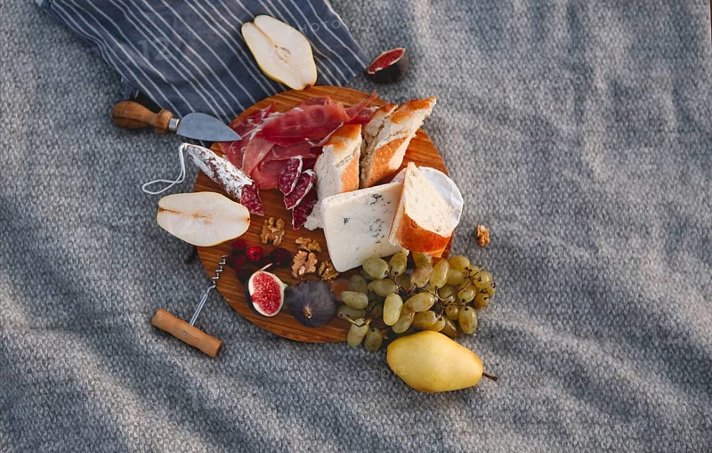 Top view of wooden board with pieces of bread pear figs nuts green grape and delicious cheese on blanket