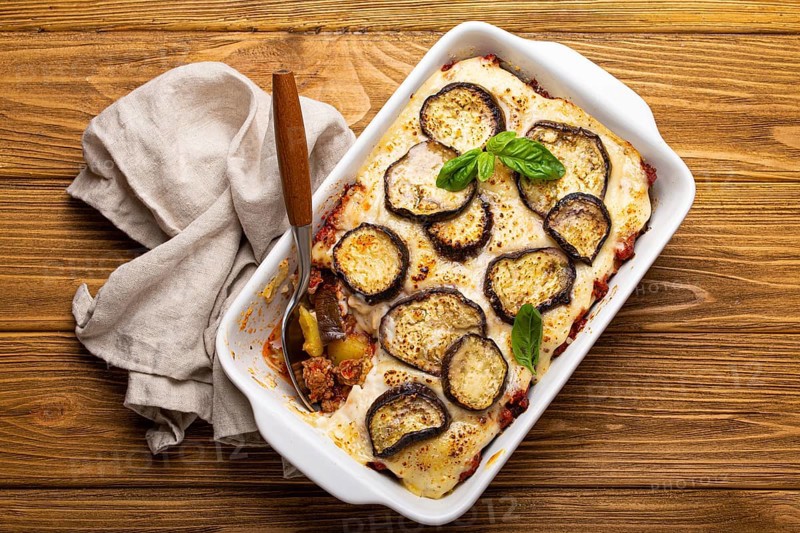 Greek mediterranean dish Moussaka with baked eggplants, ground beef in white ceramic casserole with napkin on wooden rustic background from above, traditional dish of Greece