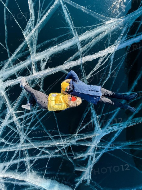 Happy couple lying on the frozen winter Lake Baikal wearing winter clothes. Transparent ice. Travel in winter, active recreation, sports, holiday