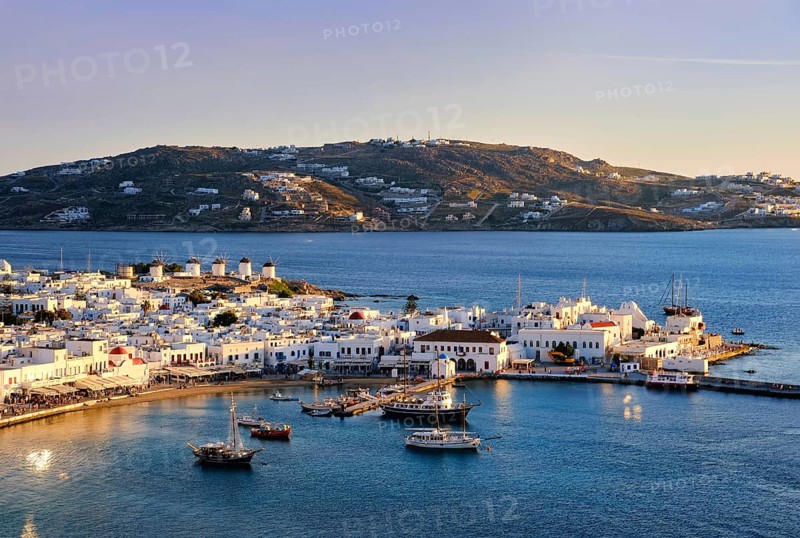 Beautiful sunset view over Chora, main town of Mykonos, Cyclades, Greece and its harbor and port. Golden hour, harbor, cruises, ship, whitewashed houses and distant islands in hazy background. Vacations, leisure, Mediterranean lifestyle