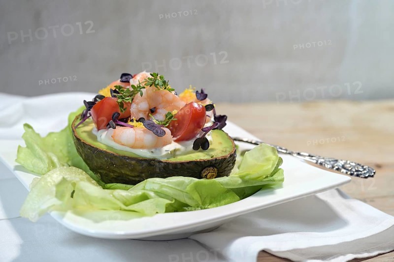 Filled avocado with shrimps, tomatoes, mayonnaise and cress garnish on lettuce and a white plate, light rustic background, festive appetizer or holiday snack, copy space, selected focus