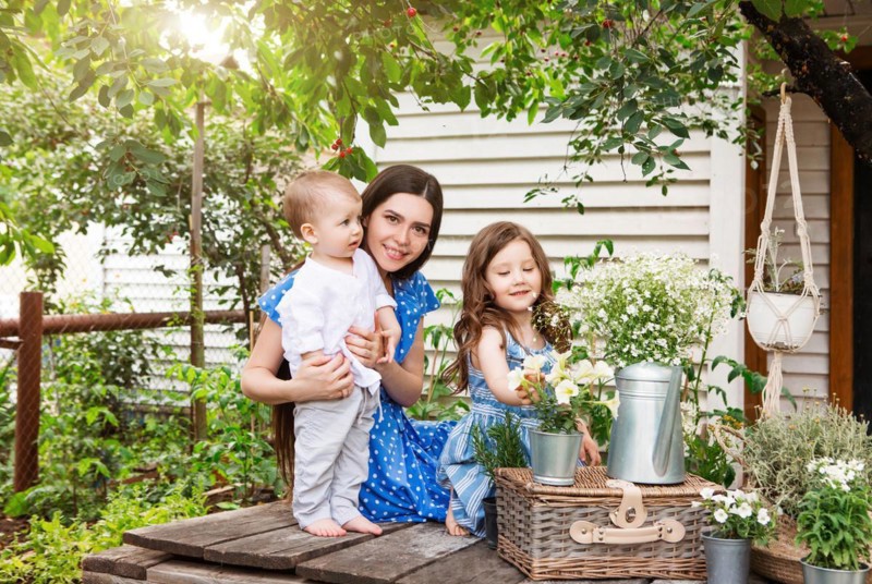 Young delighted mother sitting in armchair with adorable little siblings on terrace of country house and relaxing together during weekend