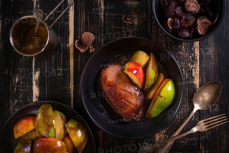 Roasted duck breast with golden crispy skin served with baked apples and chestnuts. Served on a ceramic black plate over rustic dark wooden table. Top view