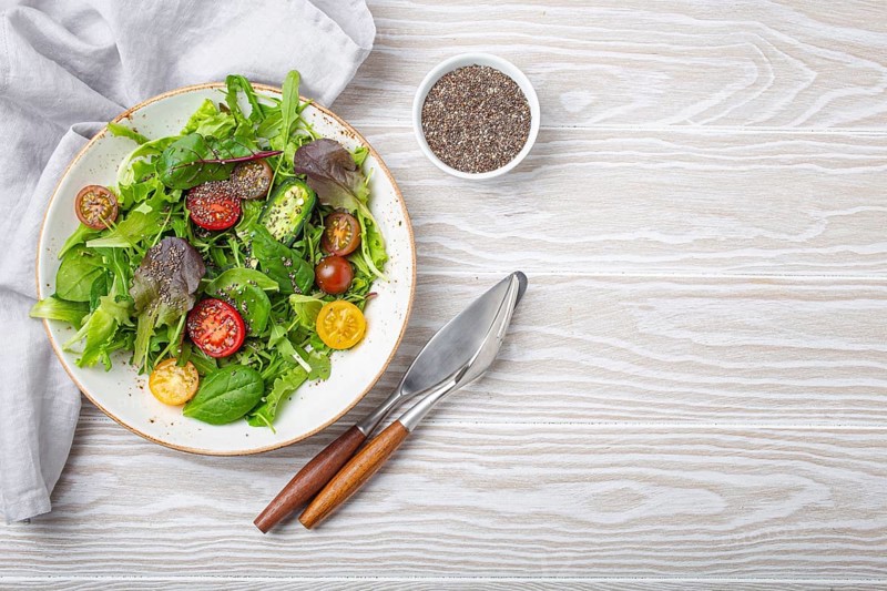 Vegetables vegetarian healthy salad with red yellow cherry tomatoes, green leafs and chia seeds on white plate on white wooden rustic background top view, healthy food and diet concept, space for text