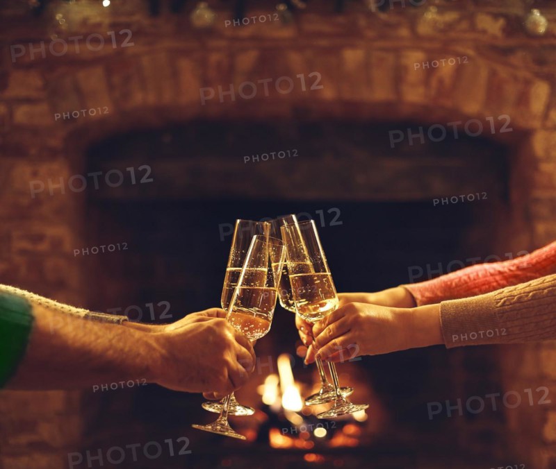 People clinking champagne glasses while gathering around fireplace in country house, cropped shot. Friends drinking alcohol, celebrating event near fire at home. selective focus. Celebration concept
