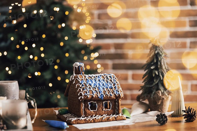 Little homemade gingerbread house standing on table with decorations, candles in living room decorated with defocused New Year lights, Christmas fir tree. Wonderful happy holiday mood. Copy space